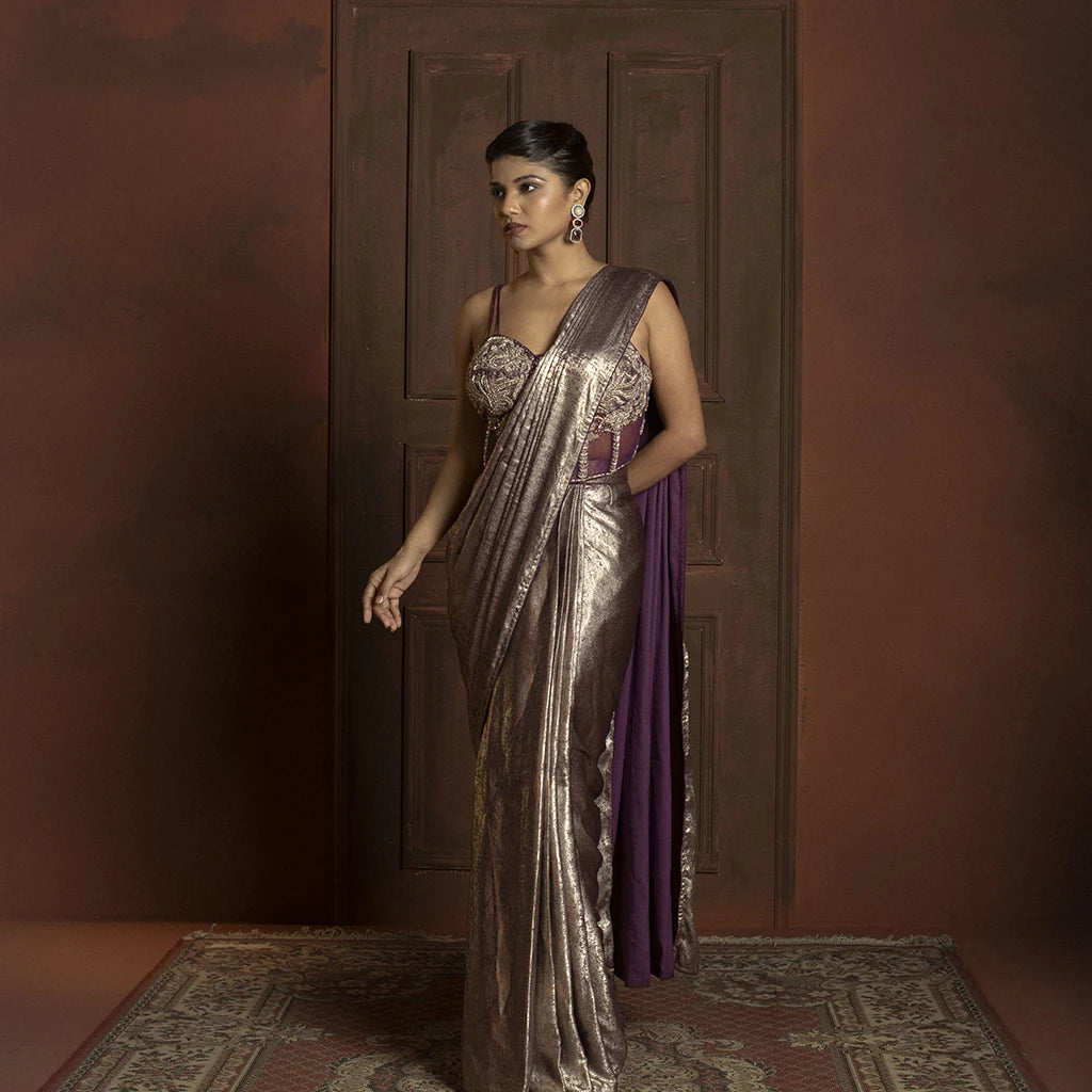 Woman in a metallic saree standing against a brown wall.