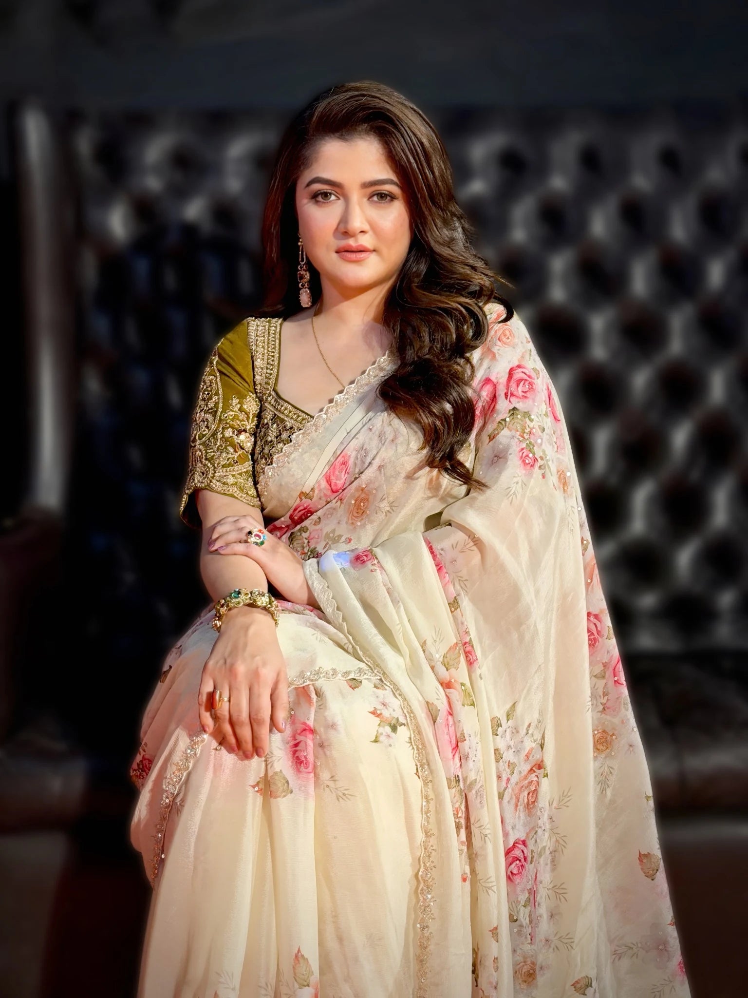 Woman in a floral saree sitting against a dark background