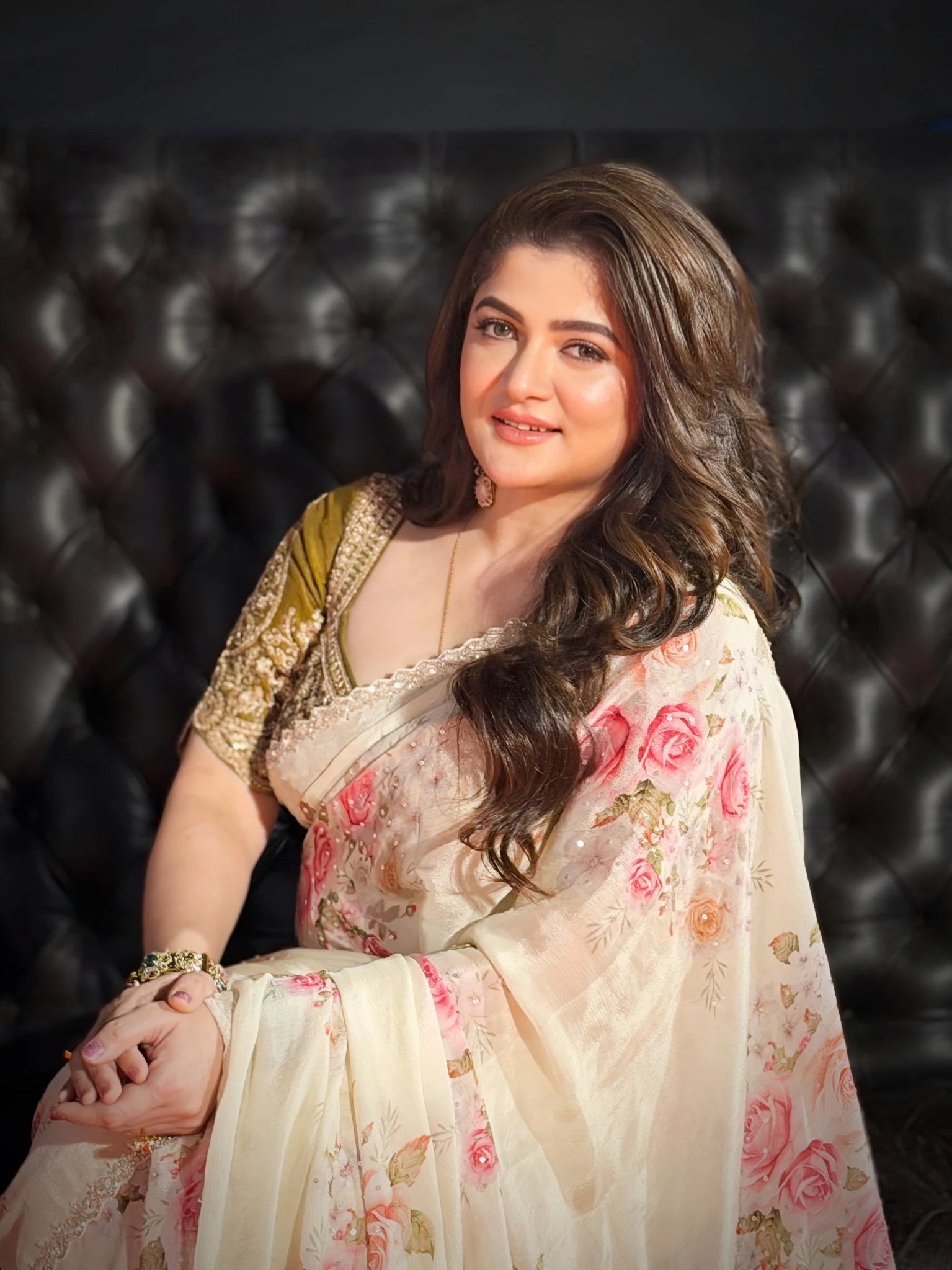 Woman in a floral saree sitting against a dark upholstered background