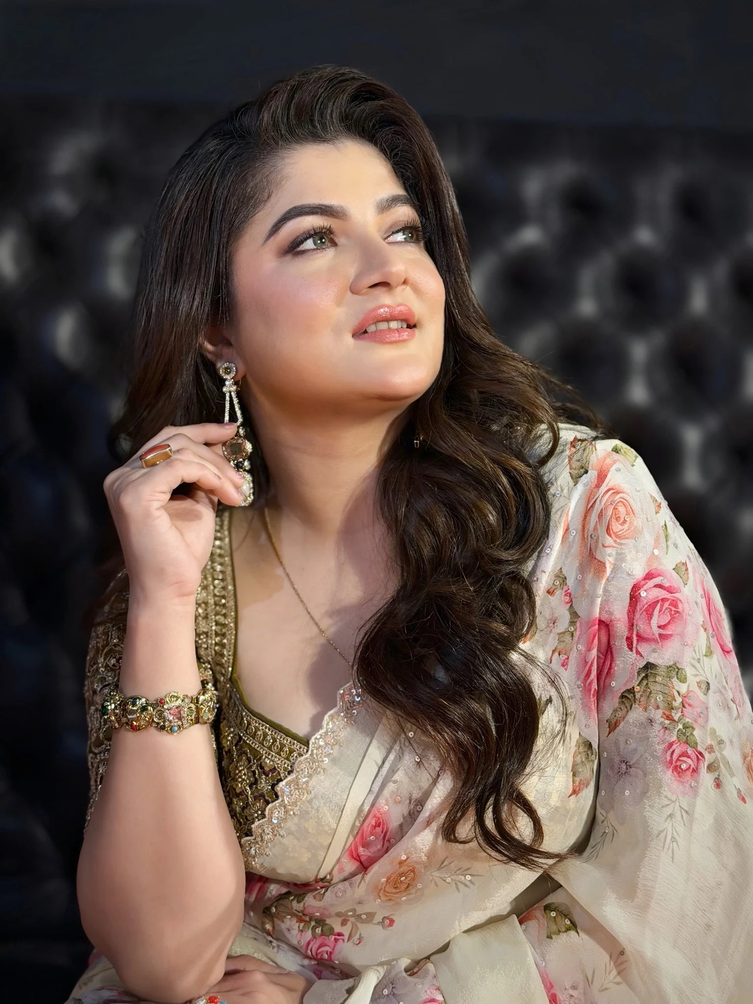Woman in a floral saree adjusting her earring against a dark background