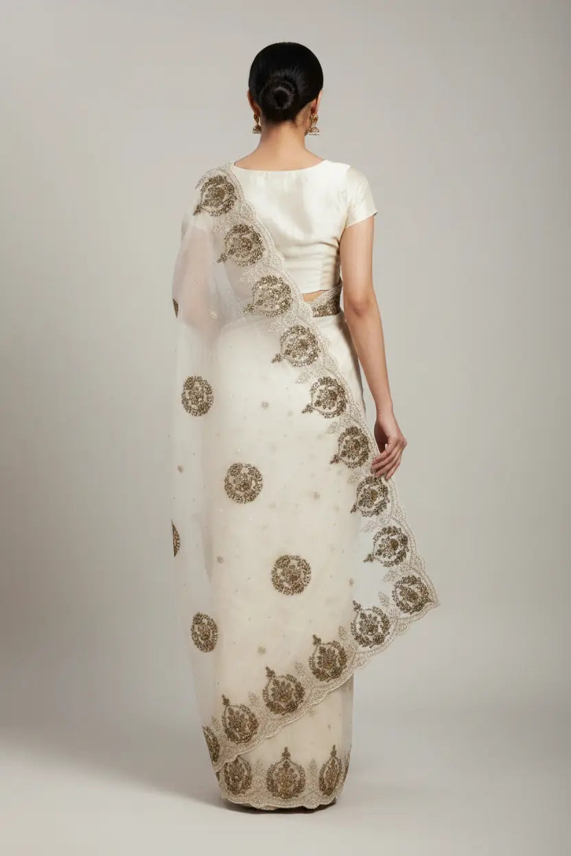 Woman wearing a white saree with gold embroidery on a plain background