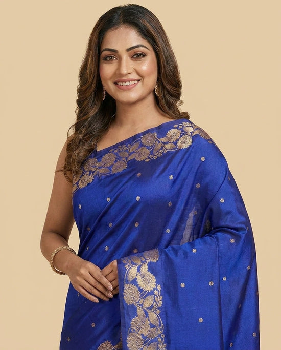 A woman wearing a royal blue silk saree with gold floral zari embroidery and small gold dots, posed against a solid beige background.