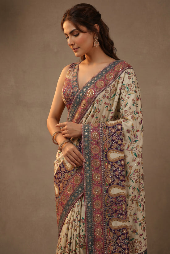 Rangoli cream silk saree with intricate purple, pink, and green paisley and floral embroidery. Features a detailed border with mirror work.