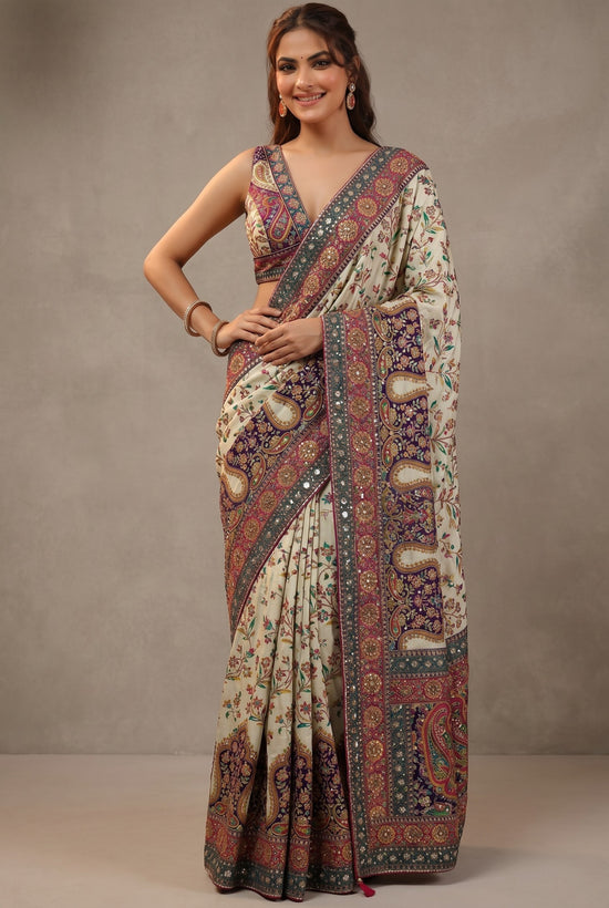 Rangoli cream silk saree with purple, pink, and green paisley and floral embroidery. Features mirror work border and matching blouse.