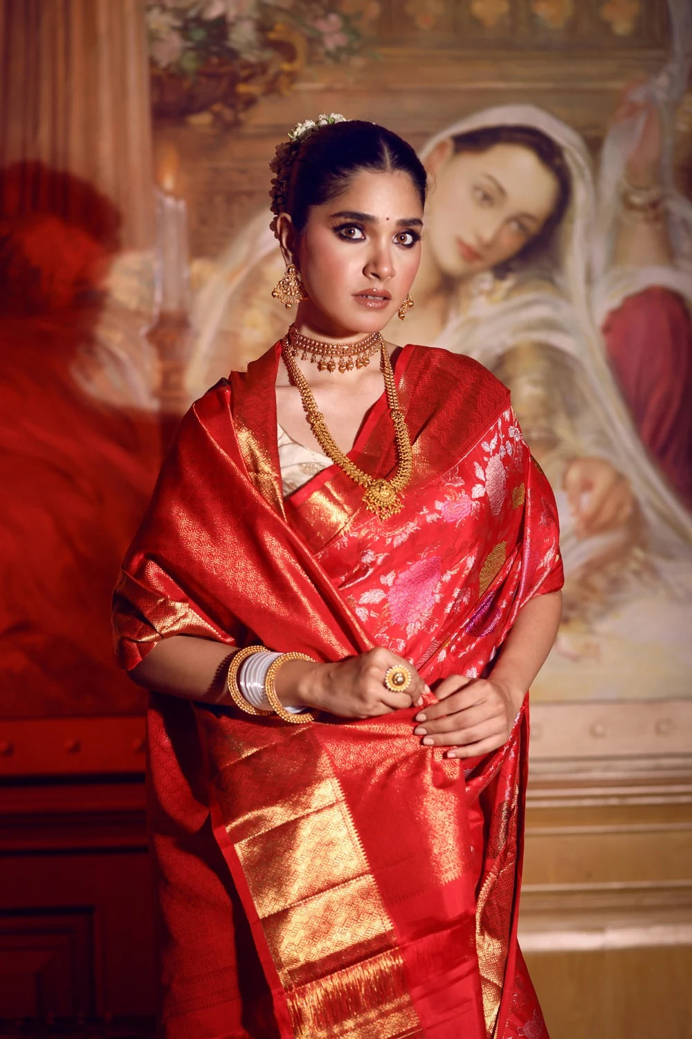 Woman in a red saree with gold details standing in front of a classical painting.