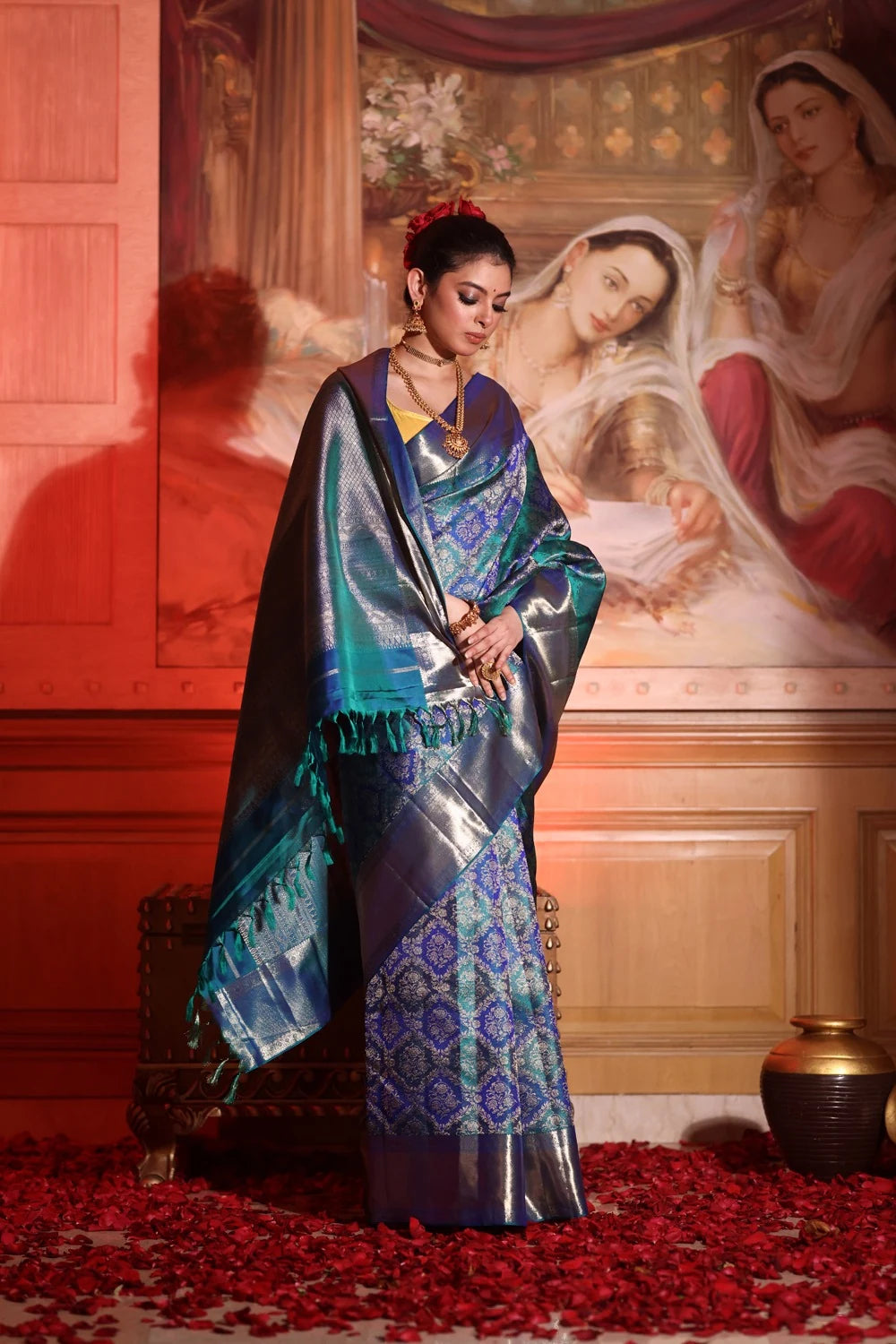 Woman in a blue saree standing in front of a classical painting with red flowers on the floor.
