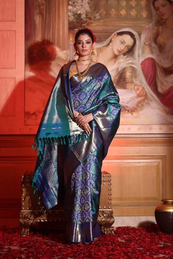 Woman in a blue saree standing in front of a classical painting with red flowers on the floor.